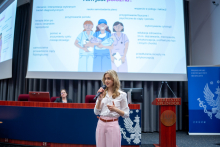  A woman stands on stage and speaks to the audience while holding a microphone; a presentation with an illustration related to medical professions is displayed behind her.