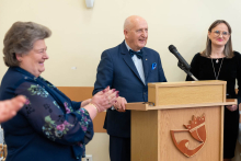 A person standing at a lectern during a ceremony, with two other people standing beside them; applause is visible in the foreground.