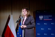 A woman standing at a microphone, dressed in a dark jacket and a light-colored blouse. The woman has her hands clasped in front of her. In the background there is a navy-blue backdrop with the coat of arms and the inscription “Ministry of Health” and the flags of Poland and the European Union.