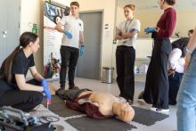 A group of participants stands around a medical manikin lying on the floor, with one person holding defibrillator pads and others observing.