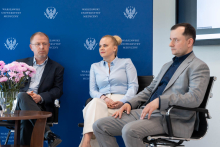 Three people sit on chairs placed side by side on a stage or in the front area of the room. In front of them stands a low table with a vase of pink flowers. In the background, there is a blue backdrop with the emblem and the name “Medical University of Warsaw,” as well as part of a presentation screen.