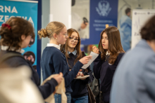 A group of young women stands at an information booth, and one of them holds a sheet of paper and reads it aloud while the others listen.