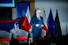 A man standing at a microphone on stage, dressed in a suit. The man is holding a folder in his hands. In the background there are armchairs, a table, and the flags of Poland and the European Union.