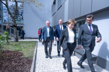 Several people walk along a paved path outside a building, dressed in formal attire, during a sunny day with trees visible nearby.