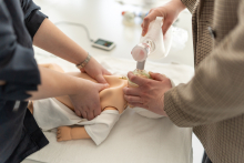 Hands hold a neonatal resuscitation mask over a baby manikin’s face during a medical training exercise.