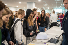 Young people gather around a demonstration table, and one person operates a device connected to a laptop placed on the table.