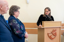 A person standing at a lectern delivers a speech during a ceremony, while two other people stand to the side listening.