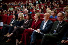 An audience seated in rows of red auditorium chairs. Women and men are looking toward the stage, and some people are holding documents or notebooks on their laps.