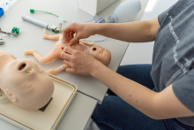 Hands manipulate a baby manikin on a table, with separate manikin parts and medical tools arranged nearby.