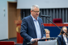 Osoba stoi przy mównicy i przemawia do mikrofonu w sali konferencyjnej. W tle widoczna jest scena z miejscami siedzącymi oraz elementy wystroju auli; na pierwszym planie pulpit z dokumentami.