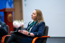 Osoba siedzi na fotelu na scenie podczas debaty panelowej i trzyma notatnik oraz długopis. Ubrana jest w elegancki strój, przed nią znajduje się stolik, a w tle widoczna jest aula konferencyjna z mównicą i elementami scenografii.