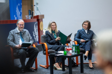 Trzy osoby siedzą na scenie podczas debaty panelowej i przeglądają dokumenty. Przed nimi znajduje się stolik z wodą i szklankami, a w tle widoczna jest mównica oraz elementy auli konferencyjnej.