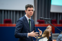 Osoba stoi przy mikrofonie na sali konferencyjnej i zabiera głos podczas debaty. Na pierwszym planie widoczne są gestykulujące dłonie oraz kartka z notatkami, w tle scena z siedzącymi uczestnikami i ekran prezentacyjny.