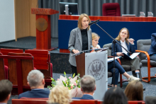 Osoba stoi przy mównicy i przemawia do mikrofonu w auli. W tle widoczni są siedzący uczestnicy debaty oraz fragment sceny z czerwonymi fotelami, a na pierwszym planie publiczność.