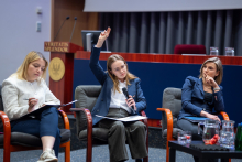 Trzy osoby siedzą na scenie podczas debaty panelowej. Jedna osoba trzyma mikrofon i podnosi rękę, pozostałe mają notatki na kolanach. Przed nimi znajduje się stolik z wodą, a w tle widoczna jest aula z mównicą i czerwonymi fotelami.