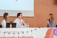 Trzy młode osoby w czasie konkursu. Kobieta siedząca po prawej mówi do mikrofonu. Dwie osoby po lewej słuchają jej. Za nimi widać ekran multimedialny,