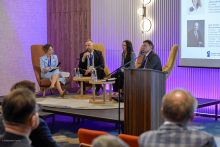 Cztery osoby: dwie kobiety i dwóch mężczyzn siedzą na scenie w czasie panelu. Jeden z mężczyzn trzyma mikrofon i przemawia. W tle widać ekran multimedialny z wyświetlanymi sylwetkami prelegentów. Widać też osoby siedzące na widowni, które słuchają dyskusji.