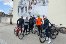 Grupa osób ubranych w stroje kolarskie i z rowerami pozuje do zdjęcia przy budynku, na ścianie którego widać mural z osobą jadącą na rowerze.