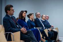 Kobiety i mężczyźni siedzą w rzędzie na krzesłach w sali konferencyjnej. Osoby mają identyfikatory na smyczach, a część z nich trzyma w rękach telefony komórkowe lub notatniki. Wszyscy skierowani są w jedną stronę, w stronę prelegentów.