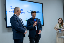 Dwóch mężczyzn stoi przed ekranem prezentacyjnym w sali konferencyjnej. Jeden z mężczyzn trzyma w ręce niebieski mikrofon i gestykuluje dłonią, drugi mężczyzna stoi obok z rękami złożonymi przed sobą. Z boku stoi kobieta trzymająca w rękach notatnik. Na ekranie wyświetlony jest napis „Konferencja Razem dla Zdrowia”.