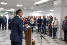 Widok od tyłu na osobę przemawiającą przy mównicy i mikrofonie. Naprzeciwko stoi liczna grupa osób słuchających przemówienia w przestronnym holu.