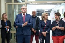 Kilka osób stoi w holu i klaszcze. Osoba znajdująca się na pierwszym planie ma dłonie złożone w geście oklasków, a inne osoby trzymają identyfikatory lub telefony.