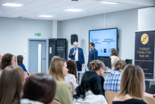 Widok z tyłu sali konferencyjnej na kobiety i mężczyzn siedzących na krzesłach. Na froncie sali dwóch mężczyzn stoi przy stole prelegenckim, a jeden z nich trzyma w ręce mikrofon. Obok znajduje się kobieta obsługująca komputer. W tle widoczny jest ekran prezentacyjny oraz baner Studenckiego Towarzystwa Naukowego.