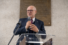 Osoba w formalnym stroju stoi przy mównicy i trzyma w dłoni mikrofon. W tle widoczna jest ściana z pamiątkową tablicą.