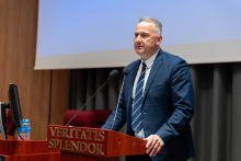 Mężczyzna stoi przy mównicy i przemawia do uczestników. Przed nią znajdują się mikrofony, a obok stoi monitor.