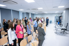 Kobiety i mężczyźni stoją przy swoich miejscach w sali konferencyjnej. Część osób trzyma w rękach torby lub ma dłonie opuszczone wzdłuż ciała. Na froncie sali znajduje się mężczyzna stojący przy mównicy oraz ekran prezentacyjny.