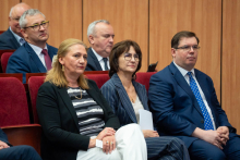 Kilka osób siedzi w rzędach na sali audytoryjnej i słucha wystąpienia. Część osób trzyma dokumenty lub programy wydarzenia.