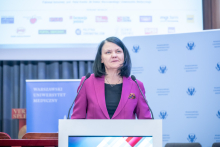 Kobieta, wiceminister zdrowia, stoi za mównicą i przemawia do mikrofonów. Za nią widoczny jest baner instytucji oraz ekran z prezentacją. Stół konferencyjny znajduje się z boku sceny.