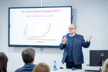 Mężczyzna stoi przed ekranem prezentacyjnym i trzyma w ręce niebieski mikrofon. Drugą ręką wykonuje gest w stronę publiczności. Na ekranie wyświetlony jest wykres oraz tekst dotyczący masy ciała i nieskutecznego odchudzania. Przed mężczyzną siedzą kobiety i mężczyźni, zwróceni w jego stronę.