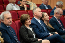 stoi przy mównicy i wygłasza prelekcję. Przed osobą znajdują się dwa mikrofony oraz pulpit z nazwą miejsca.