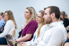 Kobiety i mężczyzna siedzą w rzędzie na krzesłach i słuchają wystąpienia. Jedna kobieta trzyma na kolanach laptop, inne osoby mają dłonie złożone lub oparte na kolanach. Wszyscy skierowani są w stronę prelegenta.