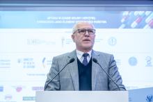 Mężczyzna, rektor WUM, stoi za mównicą i wygłasza wystąpienie. Na ekranie w tle widnieje tytuł konferencji oraz logotypy partnerów. Dwa mikrofony ustawione są przed mówcą.