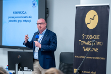 Mężczyzna stoi obok komputera i trzyma w ręce niebieski mikrofon. Drugą ręką wykonuje gest, jakby podkreślał omawiane zagadnienie. Obok znajduje się baner Studenckiego Towarzystwa Naukowego Warszawskiego Uniwersytetu Medycznego, a na ekranie wyświetlony jest slajd z hasłem „Komunikacja”.