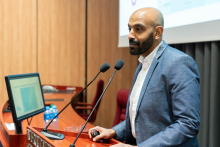 Mężczyzna stoi przy mównicy i prowadzi prezentację. Przed nią znajdują się dwa mikrofony i monitor. Osoba obsługuje pilot do slajdów.