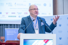 Mężczyzna, prof. Gałązkowski, stoi za mównicą i gestykuluje jedną dłonią podczas wystąpienia. Przed nią ustawione są dwa mikrofony. W tle znajdują się banery instytucji oraz ekran prezentacyjny.