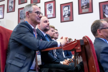 Pięć osób siedzi w pierwszym rzędzie sali wykładowej. Wszystkie patrzą w stronę sceny i słuchają wystąpienia. Na ścianie za nimi wiszą portrety dawnych przedstawicieli uczelni.