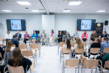 Kobiety i mężczyźni siedzą w półokręgu w dużej sali konferencyjnej. Część osób trzyma w rękach mikrofony, inne mają dłonie ułożone na kolanach. Naprzeciwko nich siedzi publiczność na krzesłach. W tle widoczne są ekrany prezentacyjne oraz baner Studenckiego Towarzystwa Naukowego.