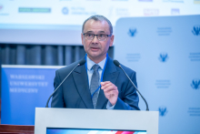 Mężczyzna, dziekan Wydziału Farmaceutycznego, stoi za mównicą i mówi do mikrofonów. Drugą dłonią wykonuje gest podkreślający wypowiedź. W tle widoczna jest ścianka z logotypami oraz fragment ekranu.