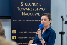 Kobieta stoi przed banerem Studenckiego Towarzystwa Naukowego i trzyma w ręce niebieski mikrofon. Drugą rękę ma opuszczoną wzdłuż ciała. Kobieta jest zwrócona w stronę publiczności i prowadzi wypowiedź.