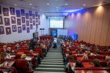 Szeroki widok sali wykładowej wypełnionej uczestnikami. Jedna osoba przemawia z mównicy na środku. Publiczność siedzi w rzędach i patrzy w stronę ekranu oraz sceny.