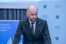 Mężczyzna, Główny Inspektor Farmaceutyczny, stoi za mównicą i przemawia do mikrofonów. Rękami podpiera się o pulpit mównicy.  Za mówcą widoczne są banery uczelni. 