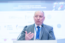 Mężczyzna, pierwszy sekretarz ambasady, stoi za mównicą i gestykuluje dłonią podczas wypowiedzi. Przed nią ustawione są dwa mikrofony. W tle widoczny jest ekran z logotypami partnerów wydarzenia.