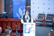 Kobieta, doradca ministra zdrowia Ukrainy, stoi za mównicą i przemawia do mikrofonów. Przed sceną siedzi publiczność. Za mówcą widoczne są banery uczelni.