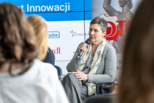 One person is seated, holding a microphone, and speaking during a panel; a graphic with a logo and part of a red inscription is visible in the background.