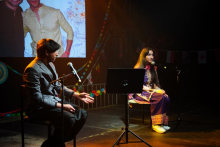 Two people seated on stage with microphones and music stands, appearing to speak or present during a live event.