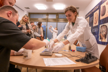 Educational workshops at a table. People wearing gloves and lab coats perform tasks using laboratory equipment and containers, while educational posters about brain structure and observing participants are visible in the background.